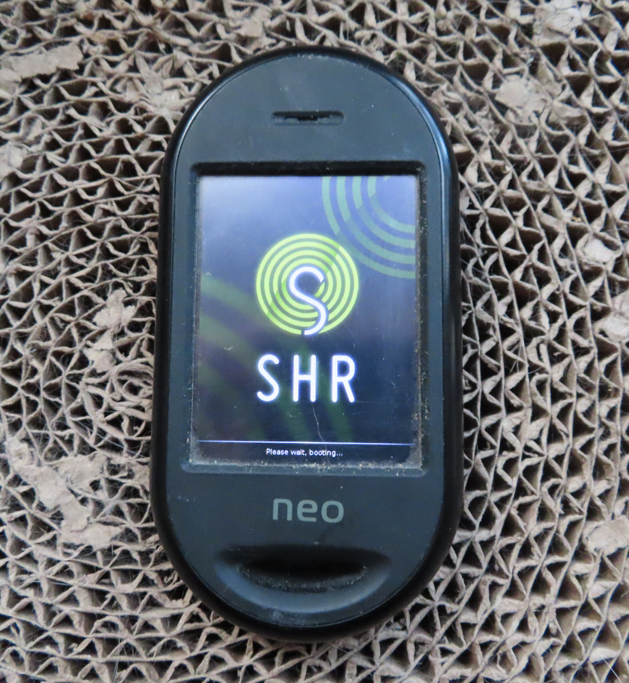 A photo of an Openmoko Neo Freerunner, displaying SHR logo and text "Please wait, booting..."